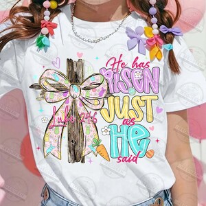 May include: White t-shirt featuring a colorful Easter design. The design includes a wooden cross with a pink bow adorned with bunnies, the text "Luke 24:6", and the phrase "He has risen just as He said" in pastel colors. Easter themed.