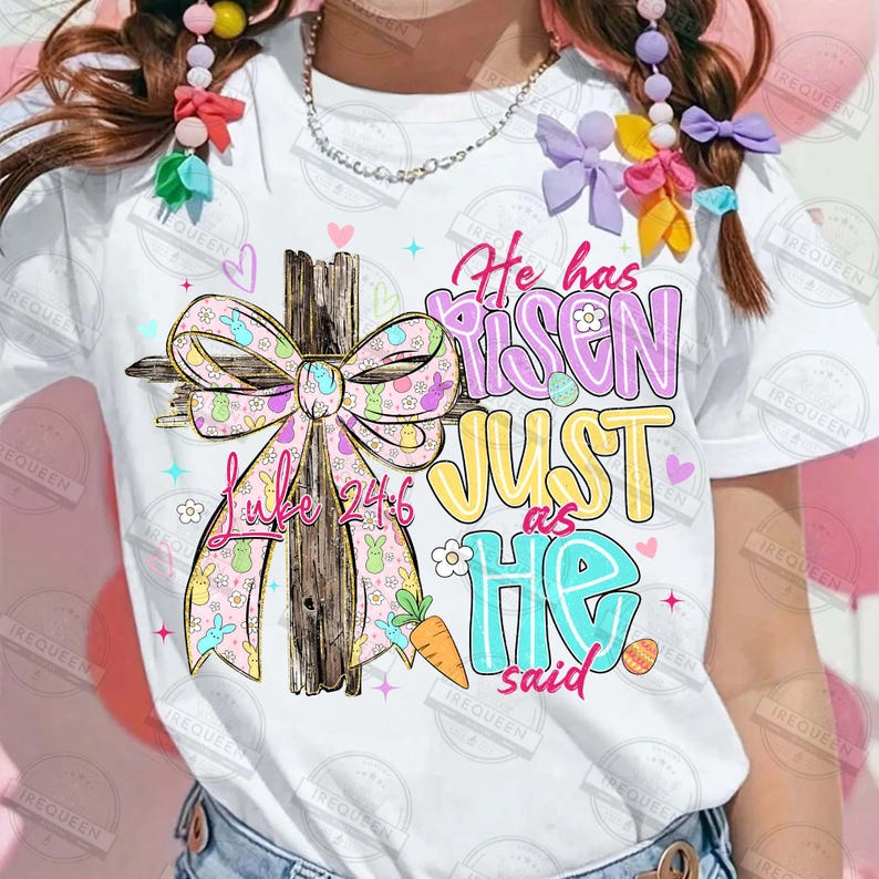 May include: White t-shirt featuring a colorful Easter design. The design includes a wooden cross with a pink bow adorned with bunnies, the text "Luke 24:6", and the phrase "He has risen just as He said" in pastel colors. Easter themed.