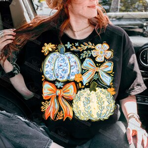May include: Black t-shirt featuring a colorful fall-themed graphic. The design includes two pumpkins with floral patterns, two bows, and various flowers in shades of orange, blue, and yellow. The shirt is a casual, everyday wear item.
