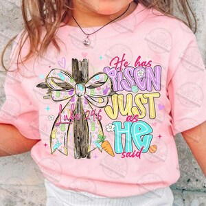 May include: Pink t-shirt with a colorful Easter-themed graphic. The design features a wooden cross adorned with a patterned bow, the text "He has Risen JUST as HE said", and the Bible verse Luke 24:6.