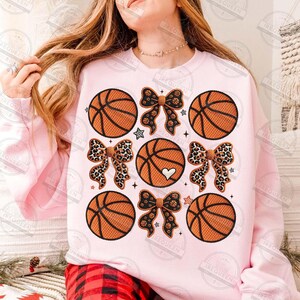 May include: A light pink sweatshirt featuring a basketball design. The design includes orange basketballs, leopard print bows, and a basketball with a white heart. The sweatshirt has long sleeves.