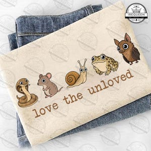 May include: A cream-colored t-shirt featuring a graphic illustration of a brown snake, a mouse, a snail, a toad, and a bat arranged in a row. Below the animals, the text "love the unloved" is printed in brown letters. The shirt is folded over blue denim.