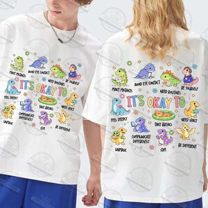 May include: White t-shirts with a colourful graphic design featuring cartoon dinosaurs and the text "It's Okay To." The design includes phrases like "Make Mistakes" and "Be Yourself."
