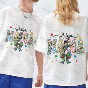 May include: Two white t-shirts with a colorful graphic design featuring the words "Autism Mama" with a puzzle piece pattern and a lightning bolt. The design includes a bow made of puzzle pieces.