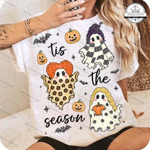 Può includere: Una t-shirt bianca con una grafica a tema Halloween e il testo "tis the season". Il design include tre fantasmi con motivi diversi, zucche arancioni e piccoli pipistrelli neri sparsi sulla parte anteriore del capo di abbigliamento.