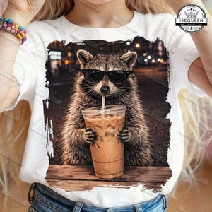 May include: White t-shirt featuring a raccoon wearing sunglasses, sipping an iced coffee. The raccoon is set against a blurred background of city lights. The shirt has the text "IREQUEEN" on it.