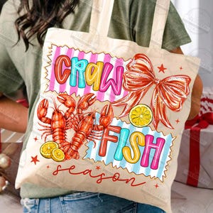 May include: A beige tote bag with a colorful design. The design includes the words "CRAWFISH" in a playful font, along with illustrations of red crawfish, lemon slices, and a red bow. The word "season" is written in a cursive font.