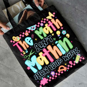 Puede incluir: Bolso tote negro con el texto "The Math Ain't Mathin' Show Your Work" en letras coloridas. El dise&ntilde;o incluye s&iacute;mbolos matem&aacute;ticos, una mariposa, flores y un l&aacute;piz. El bolso es sostenido por una persona.