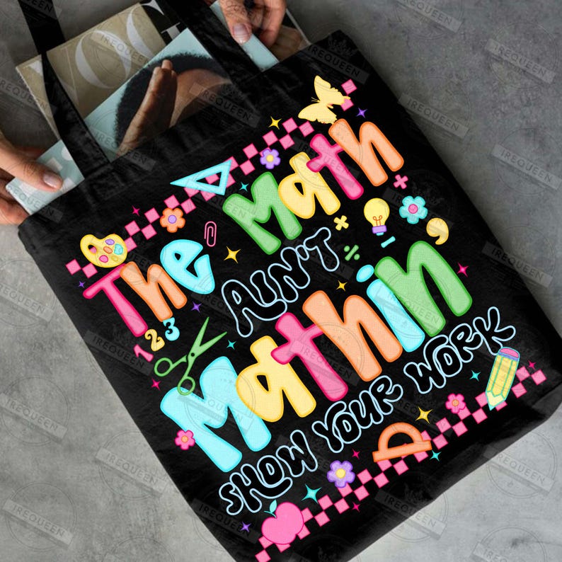 Puede incluir: Bolso tote negro con el texto "The Math Ain't Mathin' Show Your Work" en letras coloridas. El dise&ntilde;o incluye s&iacute;mbolos matem&aacute;ticos, una mariposa, flores y un l&aacute;piz. El bolso es sostenido por una persona.