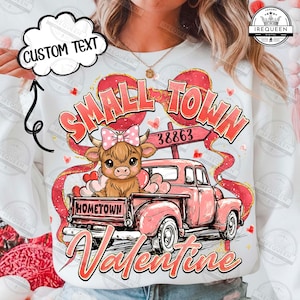 Puede incluir: Una sudadera blanca con temática de San Valentín. Presenta una camioneta rosa que transporta una vaca de las Tierras Altas con un lazo rosa, rodeada de corazones y las palabras "Small Town Valentine" y "Hometown".