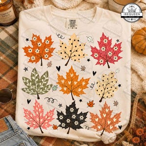 May include: A cream-colored t-shirt featuring a repeating pattern of various maple leaves in shades of orange, red, green, black, and beige. The leaves are decorated with small white flower or heart motifs and black dots. The shirt has a crew neckline.