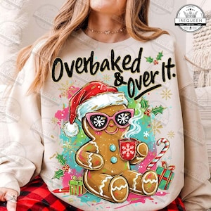 May include: A cream-colored sweatshirt featuring a gingerbread man wearing pink sunglasses and a Santa hat. The gingerbread man is holding a mug with a snowflake design. The text "Overbaked & Over It" is above the gingerbread man.