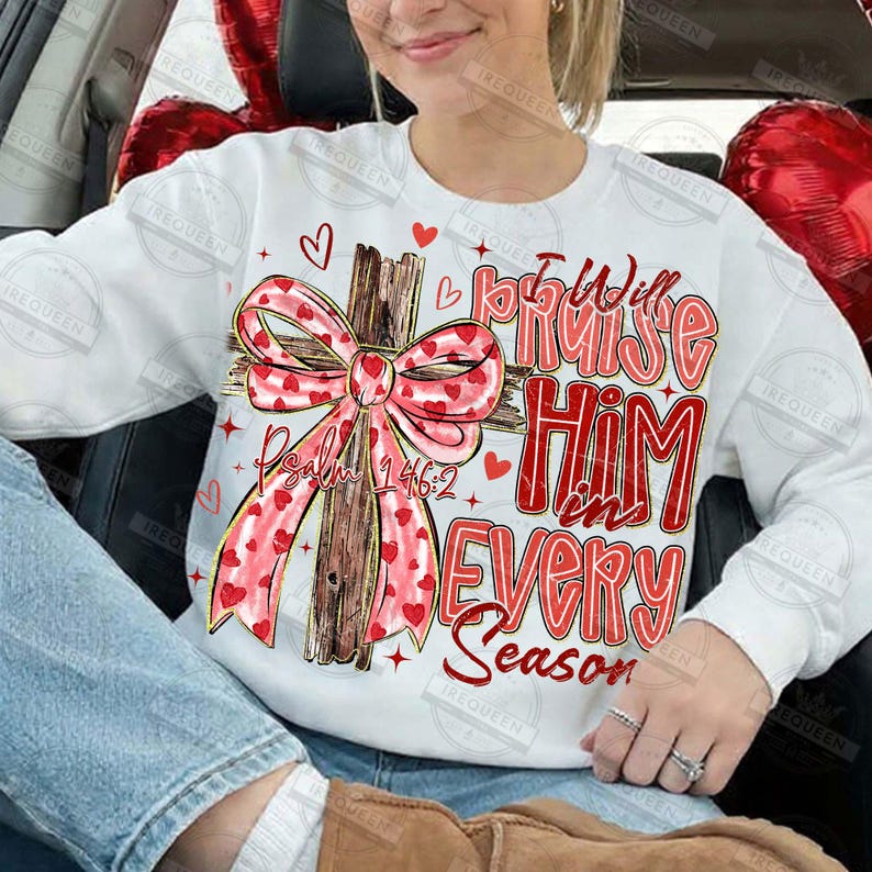 May include: White sweatshirt with a cross design. The cross is adorned with a pink bow patterned with red hearts. The text reads "I will praise Him in every season" and "Psalm 146:2" in red.