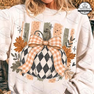 May include: A cream-colored sweatshirt featuring a central graphic of a pumpkin with a black and white diamond pattern, adorned with a large orange and white gingham bow. The design includes orange autumn leaves, foliage, and vertical stripes.