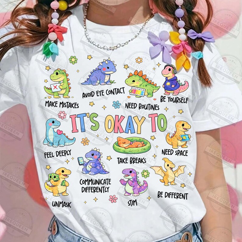 May include: White t-shirt with cartoon dinosaurs and phrases such as "Avoid Eye Contact" and "Be Yourself." The design incorporates stars and flowers, promoting a positive message. The text "It's Okay To" is also visible.