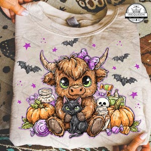 May include: A beige sweatshirt featuring a cartoon illustration of a Highland cow with a purple bow, holding a black cat. The design includes pumpkins, bats, stars, and Halloween-themed candies. The sweatshirt is folded.