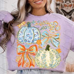 May include: A lilac-colored sweatshirt featuring a fall-themed design. The design includes two pumpkins, two bows, and floral accents in shades of blue, orange, and yellow. The pumpkins have intricate patterns, and the bows have a layered appearance.