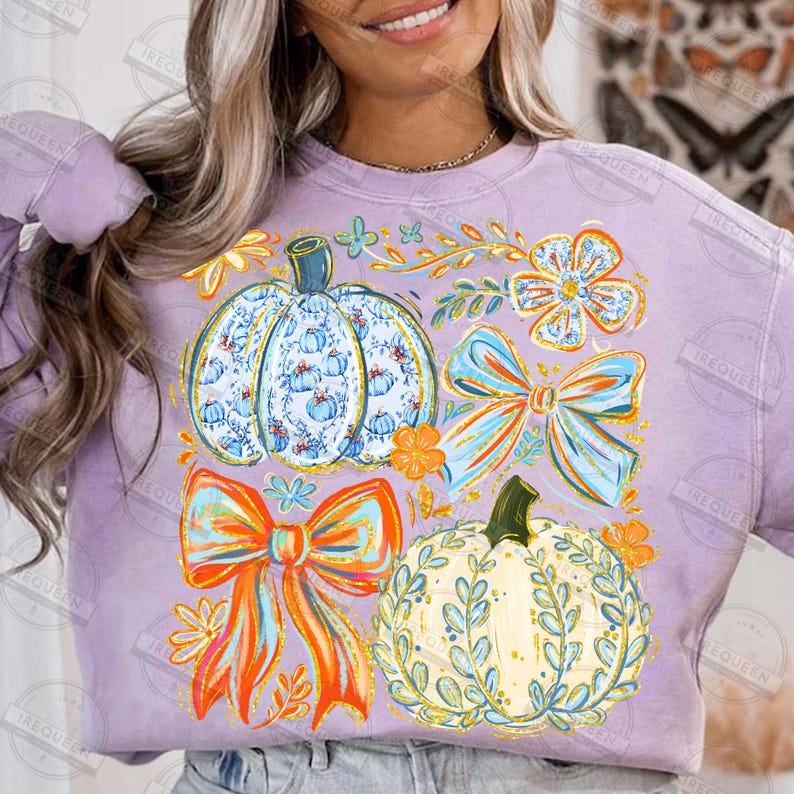 May include: A lilac-colored sweatshirt featuring a fall-themed design. The design includes two pumpkins, two bows, and floral accents in shades of blue, orange, and yellow. The pumpkins have intricate patterns, and the bows have a layered appearance.