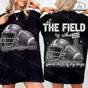May include: A black t-shirt featuring a white graphic of a football helmet and a football resting on grass. The text reads: AT THE FIELD is where I spend most of my days. The design includes a stadium light tower illustration in the background.