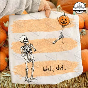 May include: A white t-shirt featuring a graphic of a skeleton looking up at a detached skeletal arm holding a floating jack-o'-lantern pumpkin balloon. The design includes three horizontal orange brush strokes with a spiderweb pattern and the text Well, shit...