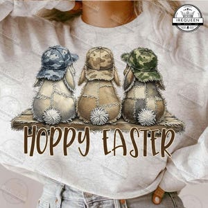 Könnte beinhalten: Drei Plüschhasenfiguren mit Patchwork-Körpern und Camouflage-Baseballkappen sitzen auf einem Holzregal. Jeder Hase hat einen flauschigen weißen Schwanz. Die Worte "HOPPY EASTER" sind darunter in brauner Schrift geschrieben.