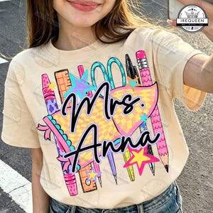 May include: A cream-colored short-sleeve t-shirt featuring a colorful graphic print of school supplies including a ruler, scissors, pencils, and crayons. The design includes a yellow checkered pennant and stars with the text Mrs. Anna in black script.