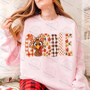 May include: A light pink sweatshirt featuring the word GOBBLE printed in large letters. The letters are decorated with orange and white plaid, autumn leaf patterns, and brown gingham. A cartoon turkey wearing a plaid bow sits in the first O.