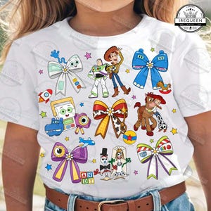 Può includere: Una t-shirt bianca con una stampa grafica colorata di vari personaggi e fiocchi. Il design include Buzz Lightyear, Woody, Jessie, Bullseye, Forky e altri elementi a tema giocattolo, intervallati da piccole stelle e un logo nell'angolo.