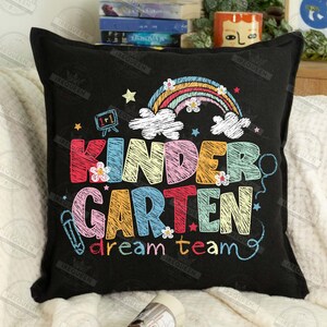 May include: Black square pillow with the words "KINDERGARTEN dream team" in colorful, whimsical lettering. The design includes a rainbow, clouds, stars, and flowers, perfect for a classroom or child's room.