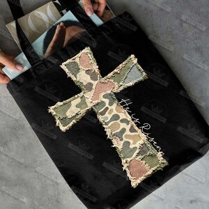 May include: Black tote bag with a large, patchwork cross design in camouflage patterns of green, brown, and tan. The words "He is Risen" are written in white script. The bag has black handles.