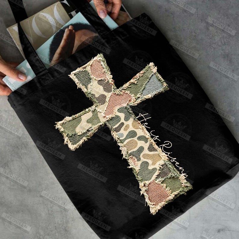 May include: Black tote bag with a large, patchwork cross design in camouflage patterns of green, brown, and tan. The words "He is Risen" are written in white script. The bag has black handles.