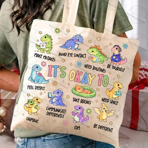 May include: A beige tote bag with dinosaur illustrations and phrases such as "Avoid Eye Contact" and "It's Okay To." The bag has a light-coloured handle. The overall theme is supportive and encouraging, with a focus on self-acceptance.