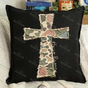May include: Black square pillow featuring a camouflage cross applique. The cross is made of patches in shades of green, brown, and beige. The words "He is Risen" are written vertically in white script on the right side of the cross.