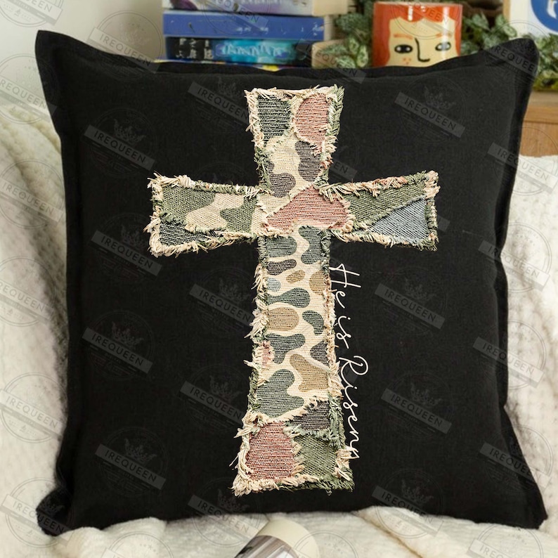 May include: Black square pillow featuring a camouflage cross applique. The cross is made of patches in shades of green, brown, and beige. The words "He is Risen" are written vertically in white script on the right side of the cross.