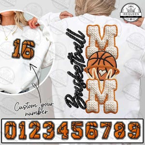 Könnte beinhalten: Weißes Sweatshirt mit dem Wort "Basketball" in schwarzer Schrift und dem Wort "MOM" in einem Strickdesign mit einer Basketballgrafik. Auf der Vorderseite befindet sich eine individuelle Nummer in Orange und Schwarz. Darunter die Zahlen 0-9.