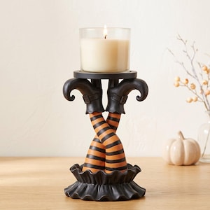 Peut inclure: Un bougeoir représentant une paire de jambes croisées portant des bas rayés orange et noirs et des bottes noires à bouts recourbés. La base est une jupe noire à volants et le sommet soutient un pot en verre contenant une bougie crème.