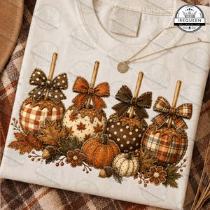 May include: A cream-colored t-shirt featuring a graphic of four caramel apples with bows, decorated with autumn leaves, acorns, and small pumpkins. The design includes plaid, polka dot, and maple leaf patterns in brown, orange, and white tones.
