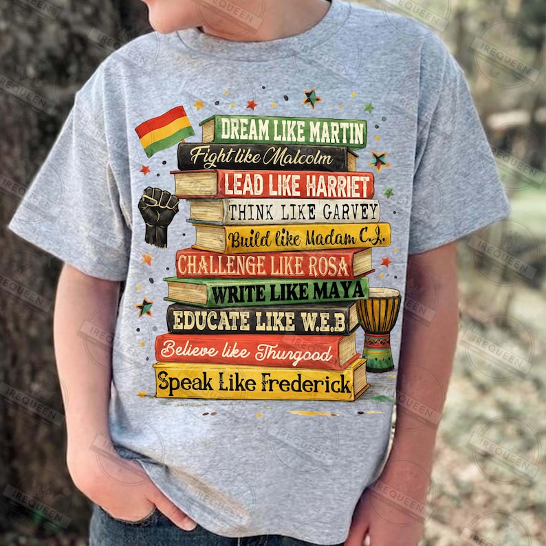 May include: A gray t-shirt featuring a stack of books with inspirational phrases like "Dream Like Martin" and "Lead Like Harriet." The design includes a black power fist, a flag, and a small drum. The shirt is worn by a person.