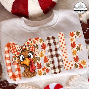 May include: A white crewneck sweatshirt featuring the text GOBBLE in large letters. The letters are filled with orange plaid, autumn leaf patterns, and brown gingham. A brown turkey illustration with a large patterned bow sits on the letter G.