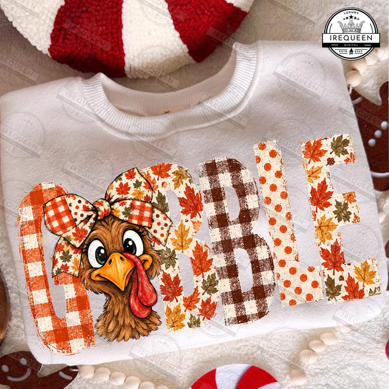 May include: A white crewneck sweatshirt featuring the text GOBBLE in large letters. The letters are filled with orange plaid, autumn leaf patterns, and brown gingham. A brown turkey illustration with a large patterned bow sits on the letter G.