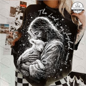 May include: Black t-shirt featuring a detailed illustration of Jesus holding a lamb, with the text "The Lord is my shepherd - Psalm 23" in white script. The design includes stars and a celestial background.