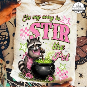 May include: A cream-colored t-shirt featuring a graphic of a raccoon wearing a pink apron and stirring a black cauldron filled with green liquid. The text reads On my way to STIR the Pot, surrounded by pink and green stars and checkered patterns.