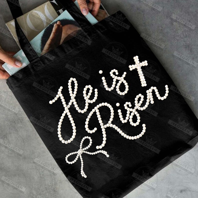 May include: Black tote bag with the phrase "He is Risen" in white, pearl-like embellishments. The design includes a cross and a bow. The bag is held open, revealing the contents inside.