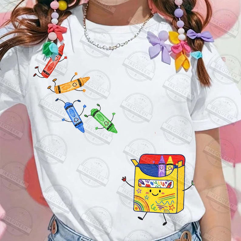 May include: A white short-sleeve t-shirt featuring a graphic print of a yellow crayon box with arms and legs, surrounded by individual red, orange, blue, and green crayon characters with faces, arms, and legs, all in a hand-drawn style.