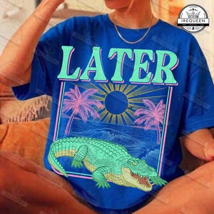 May include: Blue t-shirt with the word "LATER" in large, stylized letters. The shirt features a graphic of a beach scene with palm trees, a sun, and an alligator. The design is in shades of blue, pink, and green.