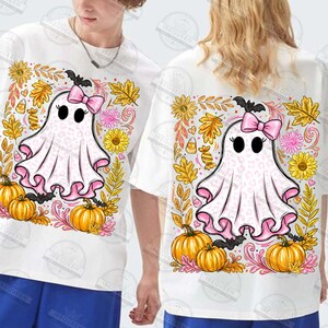 Może przedstawiać: Biały t-shirt z grafiką ducha w r&oacute;żowy wz&oacute;r panterki, noszącego r&oacute;żową kokardę. Duch jest otoczony pomarańczowymi dyniami, ż&oacute;łtymi słonecznikami, jesiennymi liśćmi i cukierkami w świątecznej, sezonowej kompozycji.