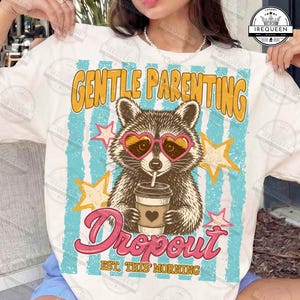 May include: White t-shirt with a raccoon graphic wearing heart-shaped sunglasses and holding a coffee cup. The shirt features the text "GENTLE PARENTING" and "Dropout EST. THIS MORNING" in a retro style.