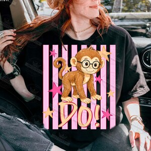 Puede incluir: Camiseta negra con un mono de dibujos animados con gafas, sobre un fondo de rayas rosas y negras. La palabra "DO!" est&aacute; escrita debajo del mono, con estrellas rosas y amarillas dispersas.