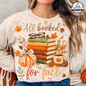 May include: A cream-colored sweatshirt featuring a graphic print of a stack of five books, a pair of glasses, a pumpkin with a patterned bow, and a mug with a maple leaf. The text reads All booked for fall, surrounded by autumn leaves and acorns.