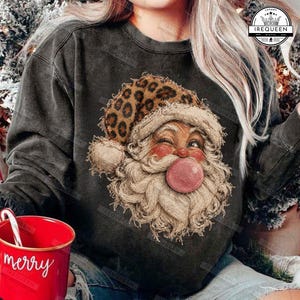 Op de afbeelding: Een antracietgrijs sweatshirt met ronde hals en een afbeelding van de Kerstman die een muts met luipaardprint draagt en een roze kauwgombel blaast. Het sweatshirt heeft een vintage gewassen afwerking en lange mouwen.